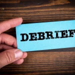 DEBRIEF concept Blue paper sheet with text in man's hands, wooden texture background